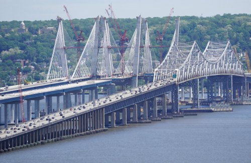 A large bridge with distinctive cables spans a river, with vehicles traveling on it and construction cranes in the background.