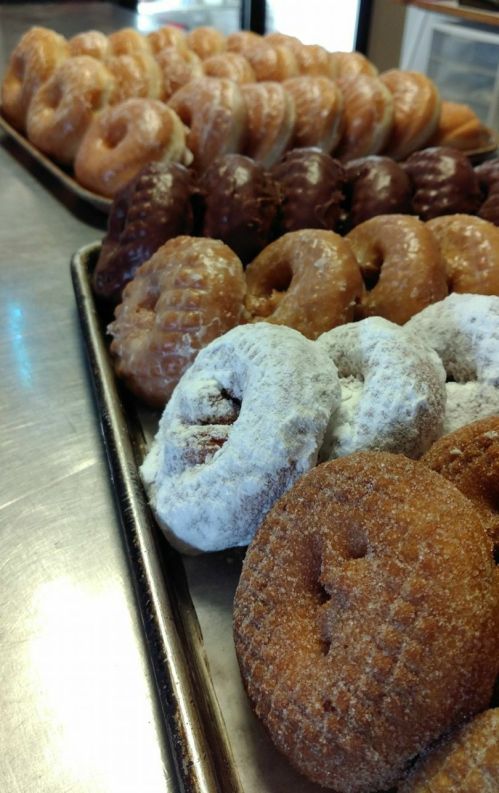 A variety of donuts on trays, including glazed, chocolate, and powdered sugar-coated options.