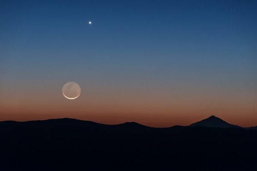 A crescent moon and a bright star shine in a twilight sky above dark mountain silhouettes.