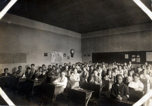 A historic classroom filled with students seated at desks, facing the camera, with a blackboard and maps on the walls.