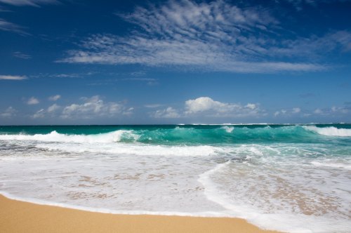 A serene beach scene with turquoise waves gently lapping at the sandy shore under a blue sky with scattered clouds.