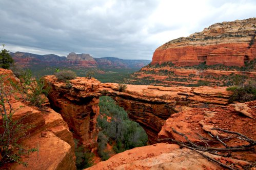 A rocky arch spans a canyon, surrounded by red rock formations and green vegetation under a cloudy sky.