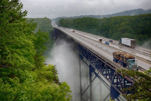 A highway bridge spans a misty valley, surrounded by lush green trees, with trucks traveling on the road.