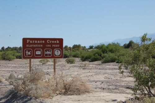 Sign for Furnace Creek, indicating an elevation of -190 feet, surrounded by desert vegetation and mountains in the background.