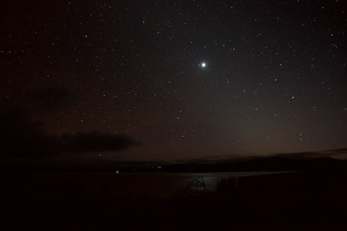 A starry night sky over a calm body of water, with a bright planet visible among the stars.