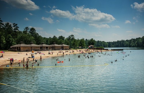A sunny beach scene with people swimming and relaxing by a lake, surrounded by trees and sandy shores.