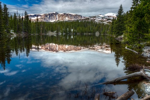 A serene lake reflects snow-capped mountains and lush pine trees under a cloudy sky.