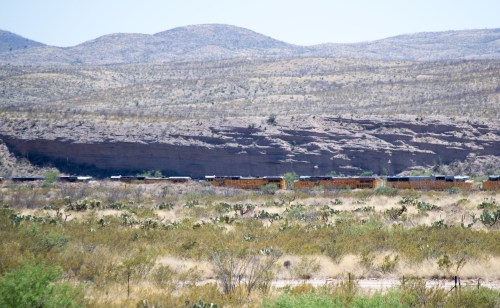 A train travels through a desert landscape with mountains in the background and sparse vegetation in the foreground.
