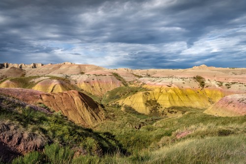 Vibrant, multicolored hills under a cloudy sky, showcasing layers of yellow, pink, and green in a natural landscape.