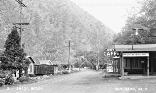 Black and white photo of a quiet road in Silverado, California, featuring a café and rustic buildings amidst hills.
