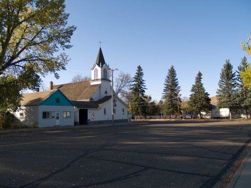 A small white church with a green trim and steeple, surrounded by trees and a clear blue sky, on a quiet street.