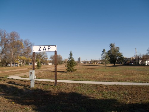 A sign reading "ZAP" stands in a grassy area with trees and buildings in the background under a clear blue sky.