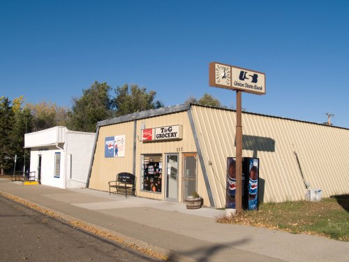 A small grocery store with a Pepsi sign and a bank sign nearby, set against a clear blue sky.