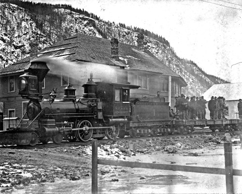 A vintage steam locomotive stops near a house, with a group of people gathered on the train's platform.