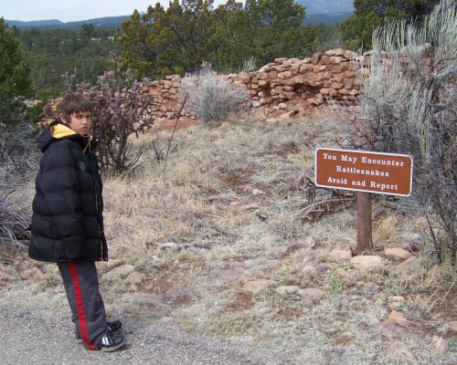 A boy in a black jacket stands near a sign warning about rattlesnakes in a rocky, wooded area.