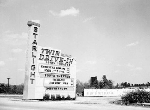 Black and white photo of the Twin Drive-In theater sign, listing movie titles and showing a vintage drive-in entrance.