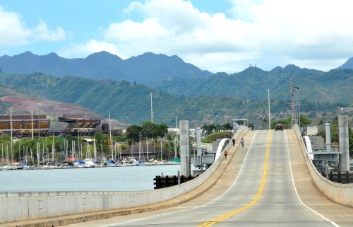 A winding road leads over a bridge with mountains and a marina in the background under a partly cloudy sky.