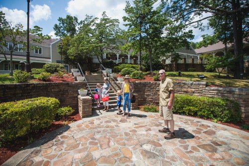 A family walks up stone steps in a park, surrounded by greenery and buildings under a blue sky.