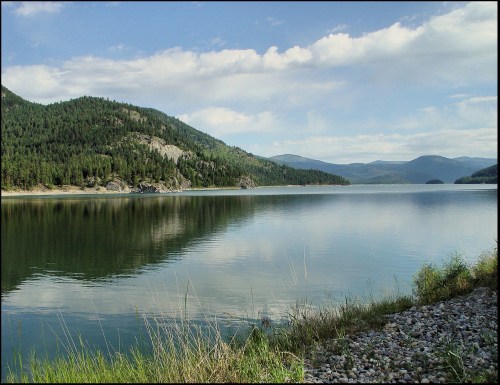 A serene lake surrounded by mountains and lush greenery, reflecting the sky and clouds on its calm surface.