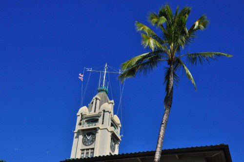 A tall clock tower with "Aloha" sign, surrounded by a palm tree against a clear blue sky.