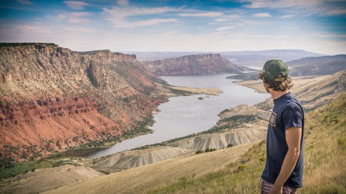A person stands on a hillside, gazing at a scenic river winding through red rock canyons under a blue sky.