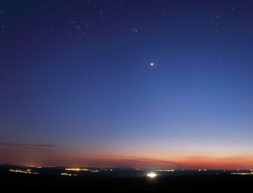 A twilight sky filled with stars, transitioning from deep blue to orange, with distant city lights visible on the horizon.