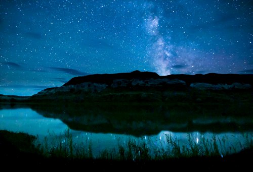 A serene night scene featuring a starry sky and the Milky Way reflected in a calm lake, surrounded by dark hills.