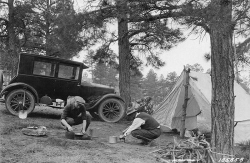 Two people cooking outdoors near a vintage car and a tent, surrounded by trees in a black-and-white photograph.