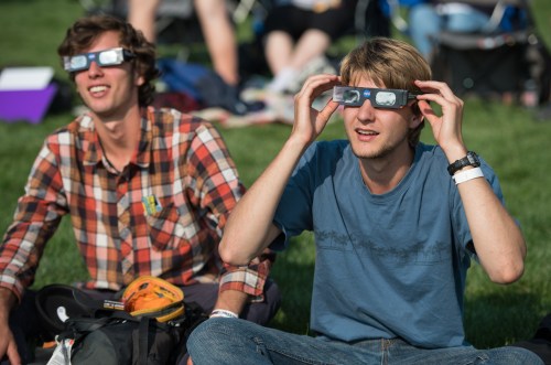 Two young men wearing eclipse glasses sit on the grass, looking up with excitement during an outdoor event.