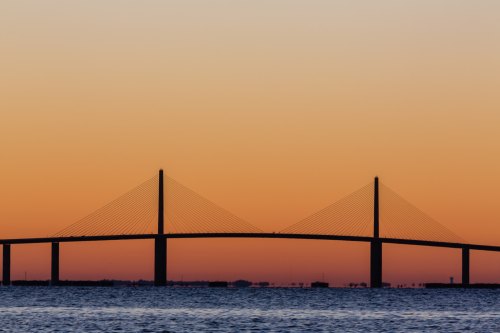 Silhouette of a bridge against a vibrant orange and blue sunset over calm waters.