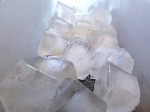 Close-up of clear ice cubes stacked in a container, reflecting light and creating a cool, refreshing appearance.