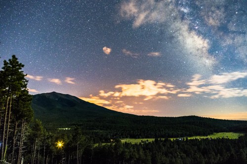 A starry night sky over a mountain, with the Milky Way visible and clouds illuminated by distant lights.