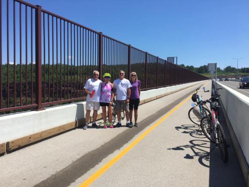 Four people stand on a bike path beside a fence, with bicycles parked nearby and a clear blue sky above.