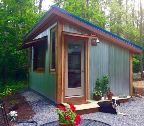 A modern, small cabin with a metal exterior, surrounded by greenery and flowers, with a dog resting on the porch.