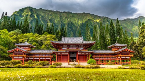 A traditional Japanese temple surrounded by lush greenery and mountains under a cloudy sky.
