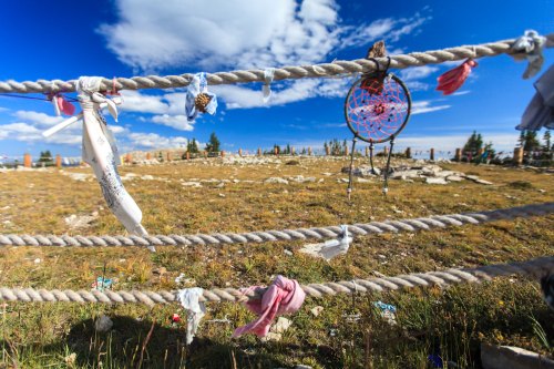 A close-up of a rope fence adorned with colorful cloth strips and a dreamcatcher, set against a blue sky and grassy landscape.