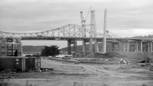 Black and white image of a bridge under construction, with cranes and piles of sand in the foreground.