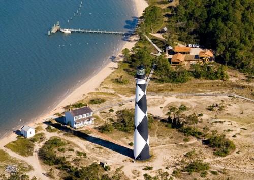 Aerial view of a black and white striped lighthouse near a sandy beach and wooden pier, surrounded by greenery.