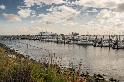 A serene marina with boats docked, surrounded by water and a cloudy sky, with a fence and greenery in the foreground.