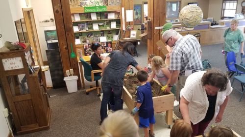 A group of children and adults engage in an interactive activity at a table filled with soil and materials in a community space.