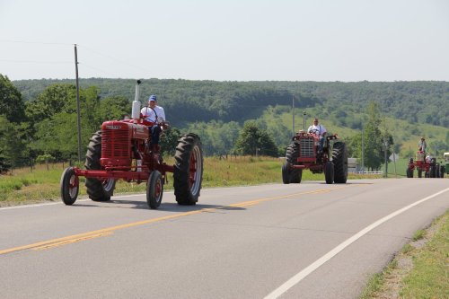 Two vintage red tractors drive down a rural road, surrounded by green hills and trees on a sunny day.