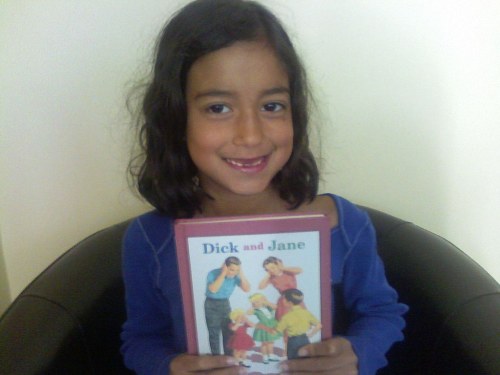 A smiling girl with dark hair holds a "Dick and Jane" book while sitting in a chair.