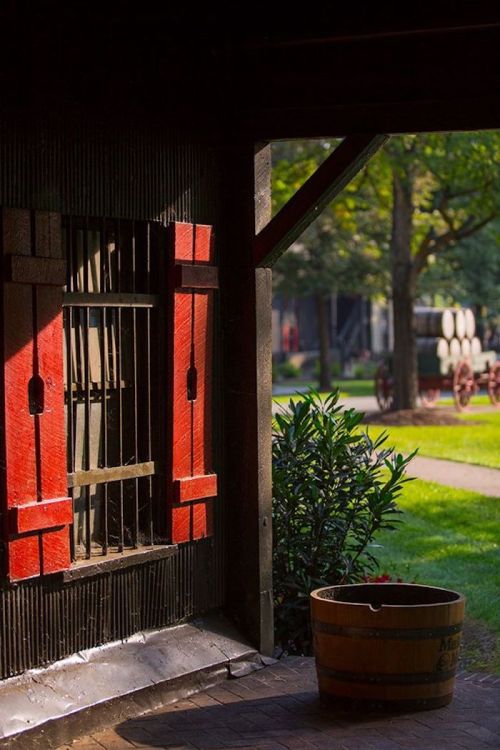 A rustic building with red shutters, a wooden barrel, and a view of a green garden and vintage wagons outside.