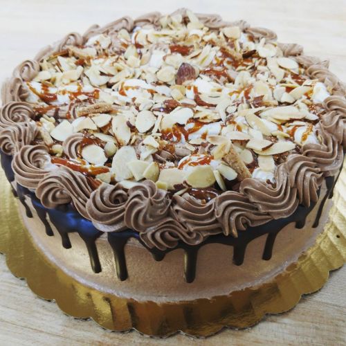 A round chocolate cake topped with caramel, sliced almonds, and decorative chocolate frosting on a gold cake board.
