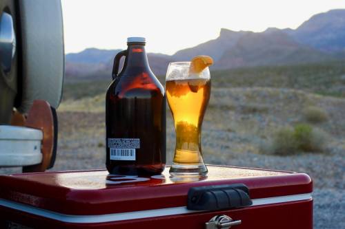 A glass of beer with an orange slice and a growler sit on a red cooler, with mountains in the background at sunset.