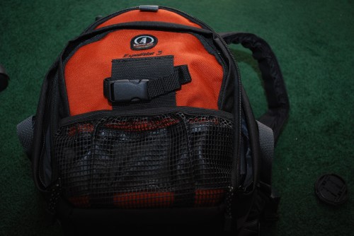 A close-up of an orange and black backpack with a mesh pocket on the front, resting on a green surface.