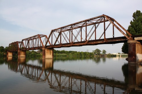 Rusty railway bridge over a calm river, reflecting in the water, surrounded by greenery and a clear sky.