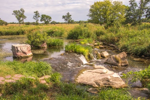 A serene landscape featuring a small waterfall over rocks, surrounded by lush greenery and a calm river.