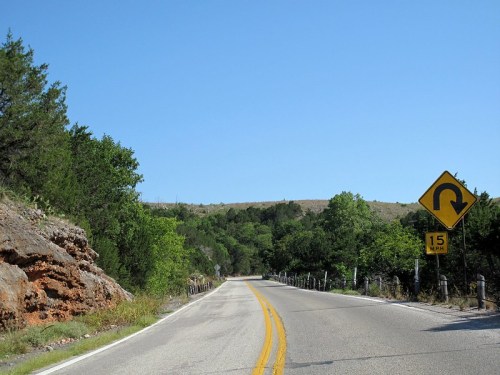 Winding road with a sharp turn sign, surrounded by greenery and rocky terrain under a clear blue sky.