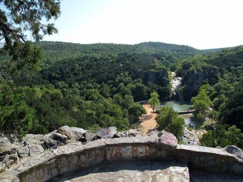 A scenic view of a lush green landscape with a waterfall and a pond, surrounded by trees under a clear blue sky.